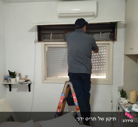 אדם עובד על תריסים בחדר עם סולם ומזגן
